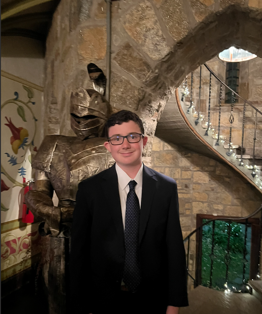 Ben Williams, founder of TerraScale Studio — a young environmental researcher and brand strategist in a dark suit and tie, photographed inside a stone-arched hall beside a suit of armor and a spiral staircase.